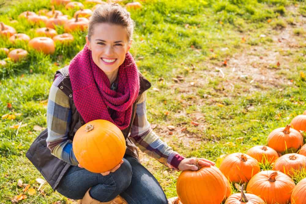 A smiling woman in a plaid shirt, vest, and chunky red scarf kneels in a field of bright orange pumpkins. Fall in Vancouver is ideal for pumpkin patch outings, where layers and rugged boots keep you comfortable and stylish on crisp afternoons.