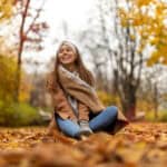 A woman in a tan coat, thick scarf, and grey gloves sits smiling on a path blanketed in fall leaves. With the trees in full color, Vancouver in autumn is perfect for cozy city strolls layered up in neutral tones.