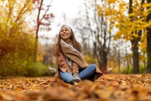 A woman in a tan coat, thick scarf, and grey gloves sits smiling on a path blanketed in fall leaves. With the trees in full color, Vancouver in autumn is perfect for cozy city strolls layered up in neutral tones.