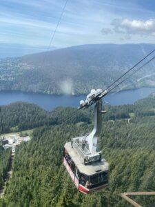 A red and white Grouse Mountain gondola glides high above dense evergreen forest, with the Capilano Reservoir and coastal mountains stretching out behind it. The ride up is as thrilling as the destination, offering panoramic views that make you feel suspended between city and wilderness.