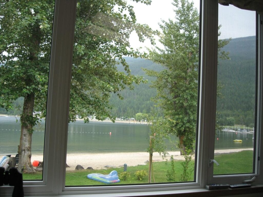 A view through a cabin window onto Mara Lake&rsquo;s sandy beach and calm waters in Salmon Arm, BC, framed by trees and mountains&mdash;showcasing one of the top things to do in Salmon Arm BC, with waterfront relaxation and scenic views.