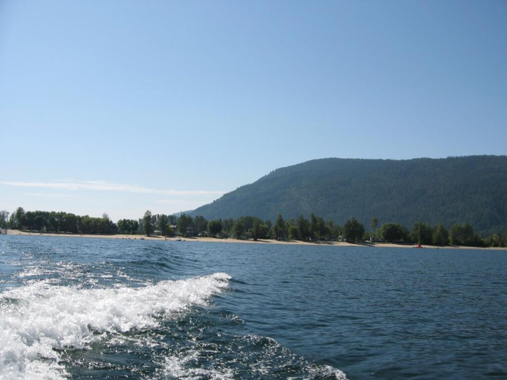 A wake racing across Mara Lake behind a small pleasure boat, with forested hills in the distance&mdash;capturing one of the most popular fun things to do in Salmon Arm BC: boating across Shuswap Lake.