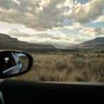 A car door is open, framing a view of rolling hills, desert-like grasses, and layered skies near Juniper Beach Provincial Park. Just outside Kamloops, the drive along the Thompson River is a quiet escape into raw, wide-open beauty.
