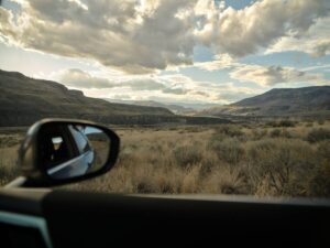 A car door is open, framing a view of rolling hills, desert-like grasses, and layered skies near Juniper Beach Provincial Park. Just outside Kamloops, the drive along the Thompson River is a quiet escape into raw, wide-open beauty.