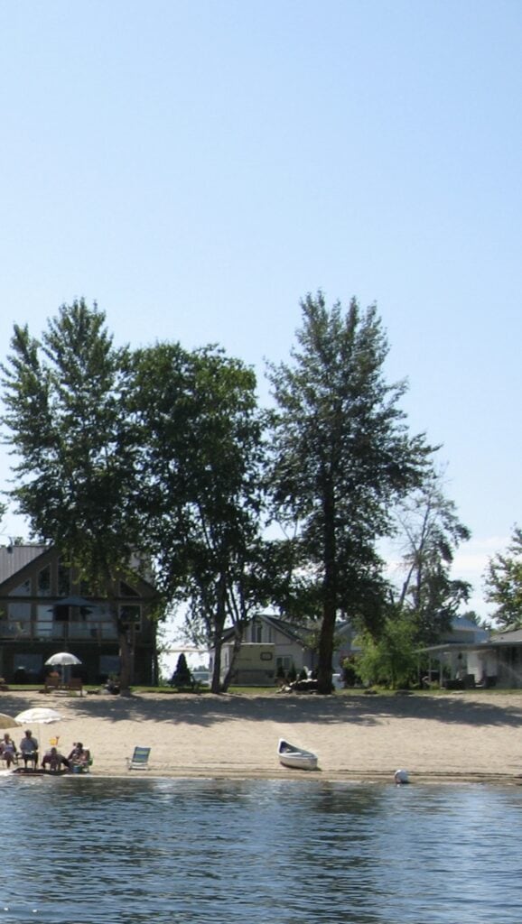 Beachfront cabins and trees line the shore of Salmon Arm while people relax in lawn chairs under umbrellas, with a canoe and beach toys nearby. A sunny summer day enjoying lakeside living in the Shuswap.