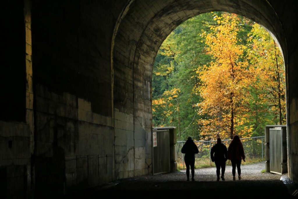 Three hikers walk out of a dark tunnel toward a glowing forest filled with golden autumn leaves. The exit frames the seasonal beauty found along this historic trail near Hope, BC.