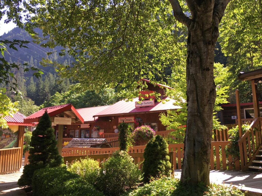 Shaded garden area with red-roofed fudge shop, ice cream stand, and signs welcoming visitors under the trees.