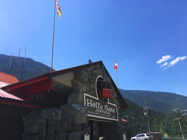Entrance to Hell’s Gate Airtram featuring a red cable car replica, BC and Canadian flags, and a backdrop of pine-covered mountains.