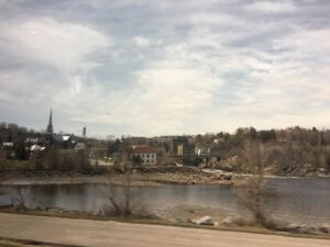 A small Quebec town sits along a riverbank, with a tall church steeple and a mix of older buildings framed by bare trees and a cloudy sky.