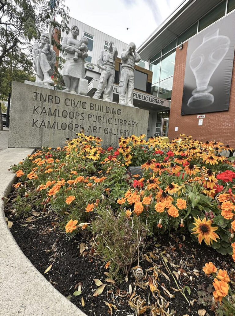 A flowerbed with vibrant orange and red flowers in front of a statue group and engraved sign for the TNRD Civic Building, Kamloops Public Library, and Kamloops Art Gallery.