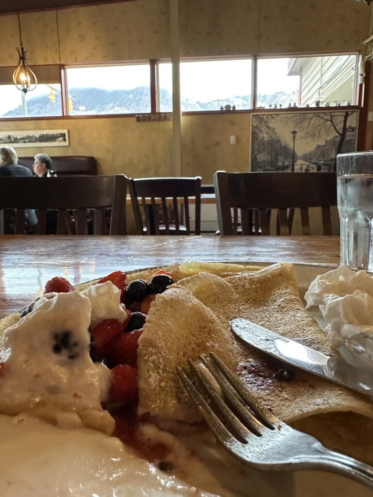 Partially eaten crepes filled with berries and whipped cream, with a fork and knife resting on the plate inside Amsterdam Restaurant with windows looking out toward the hills.