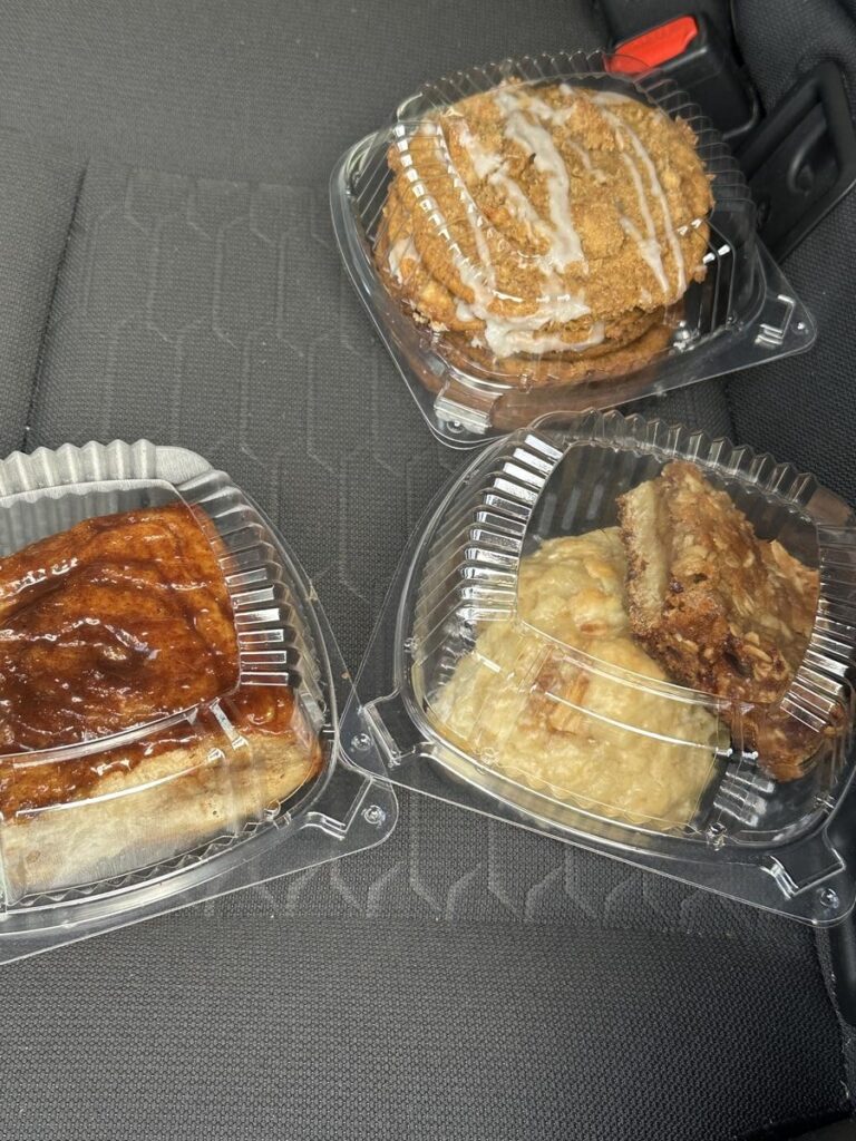 Three plastic containers of Brynn’s Bakery pastries on a car seat, including iced cookies, sticky buns, and dessert bars.