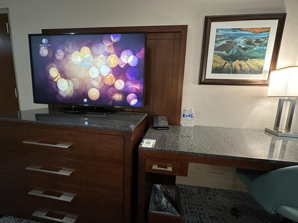 Hotel room workstation with a flat-screen TV, desk area, two water bottles, phone, and a framed abstract landscape above a lamp-lit workspace.