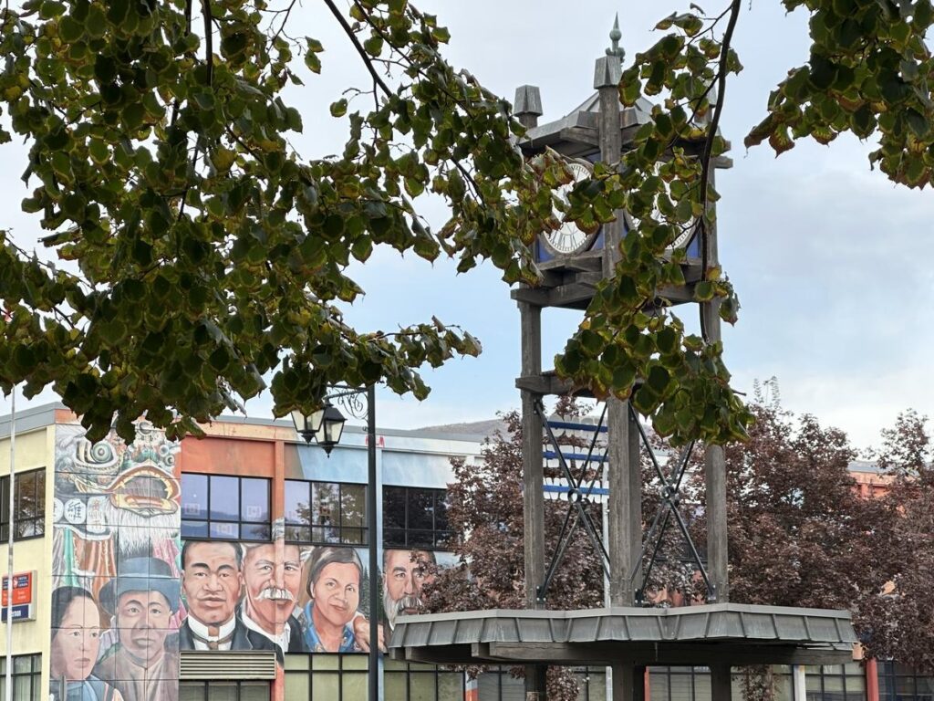 Vernon&rsquo;s downtown clock tower framed by leafy trees, with a colorful mural of historical community figures in the background.