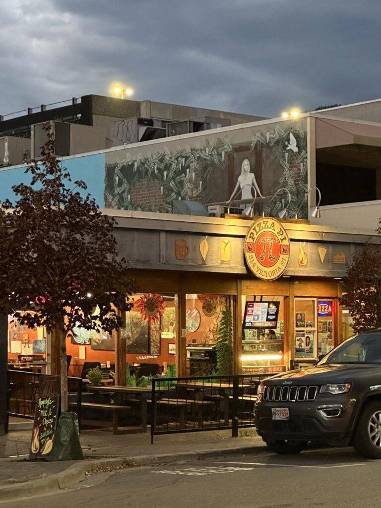 Exterior view of Pizza Pi in Kamloops at dusk with lights glowing, murals on the walls, and patio seating out front.