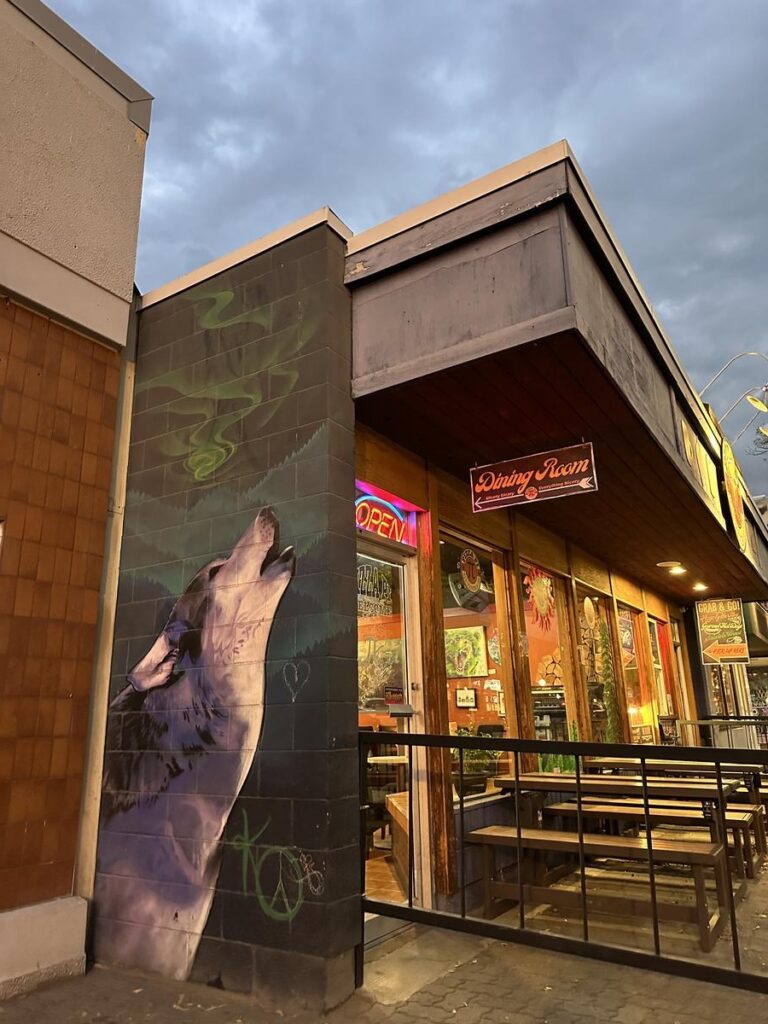 Side view of Pizza Pi&rsquo;s entrance with a neon open sign and a mural of a howling wolf on the corner wall.