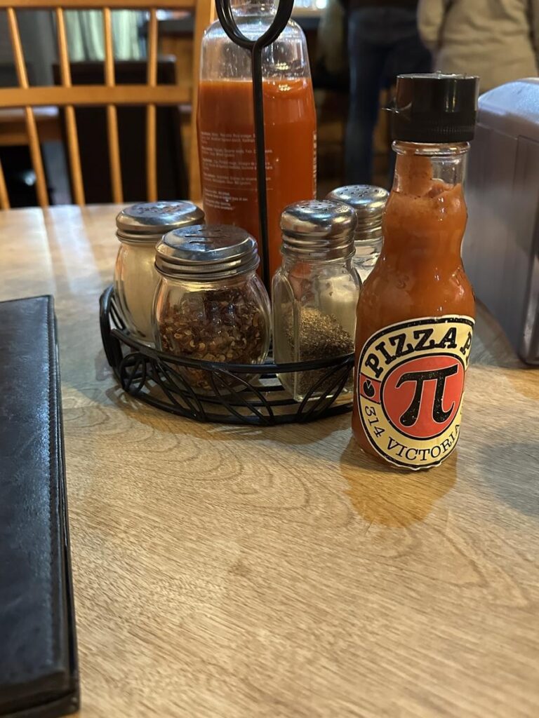Tabletop condiment set with Pizza Pi-branded hot sauce, chili flakes, pepper, and salt shakers.