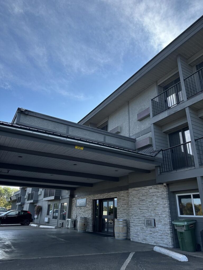 Exterior view of Quality Inn & Suites Vernon showing the covered main entrance, modern stone facade, and parking lot under a blue sky.