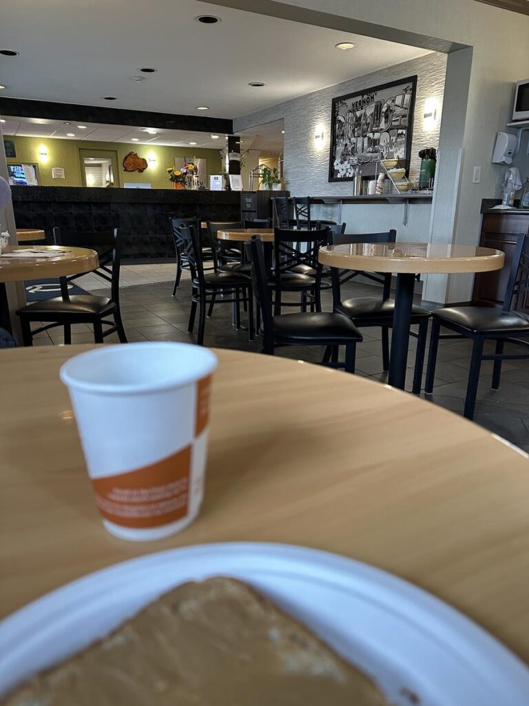 Close-up of a breakfast table with a slice of toast and a paper coffee cup in the foreground, looking toward the dining area at Quality Inn Vernon.