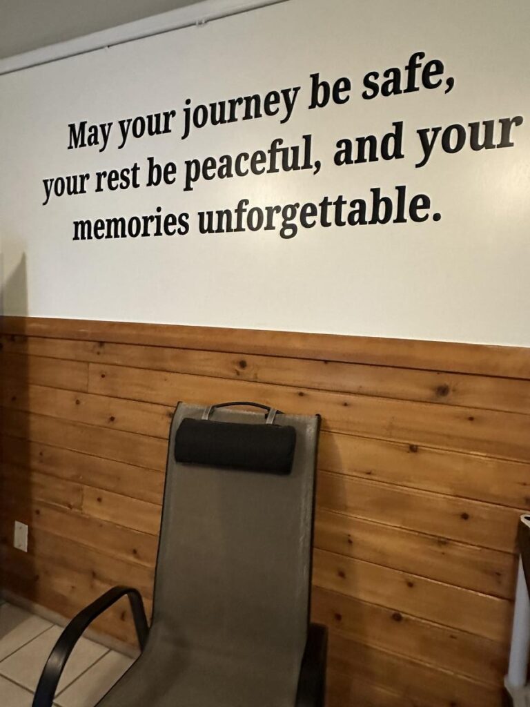 Inspirational wall quote reading &ldquo;May your journey be safe, your rest be peaceful, and your memories unforgettable&rdquo; beside a lounge chair and wood-paneled wall.