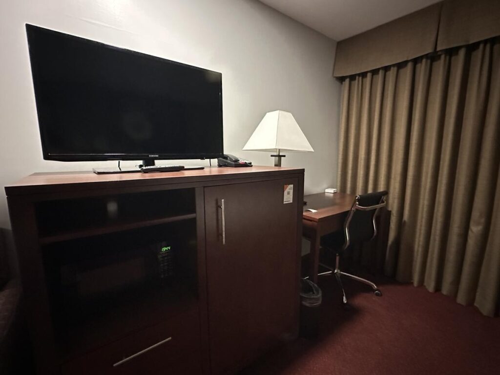 Hotel room amenities including a flat-screen TV, microwave, and mini fridge on a dark wood cabinet, next to a work desk and chair.