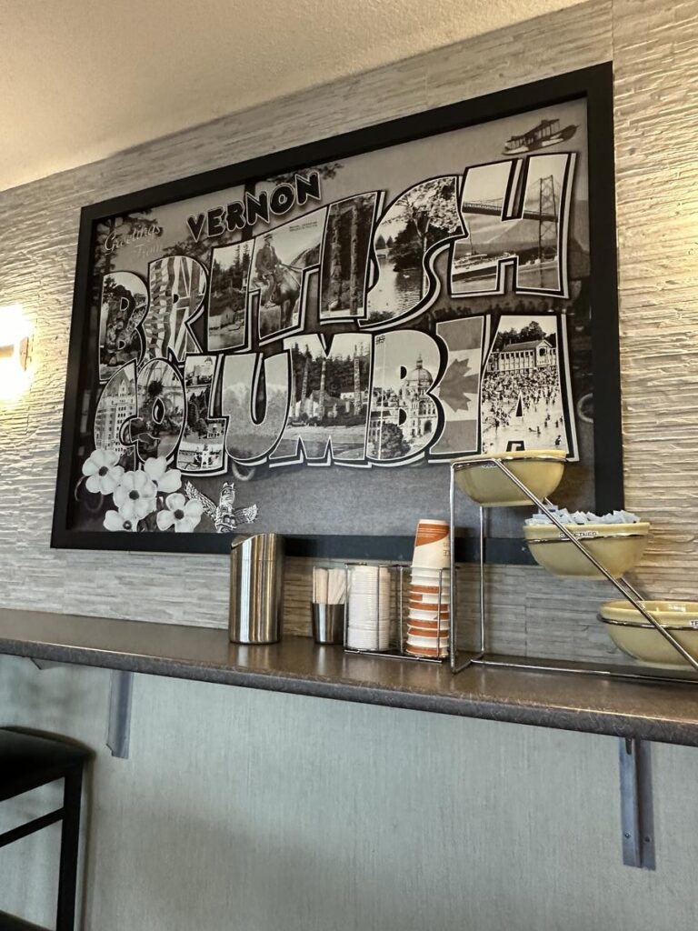 Black and white framed artwork reading "Vernon British Columbia" in large block letters filled with local scenic imagery, mounted above a coffee station with cups and lids.