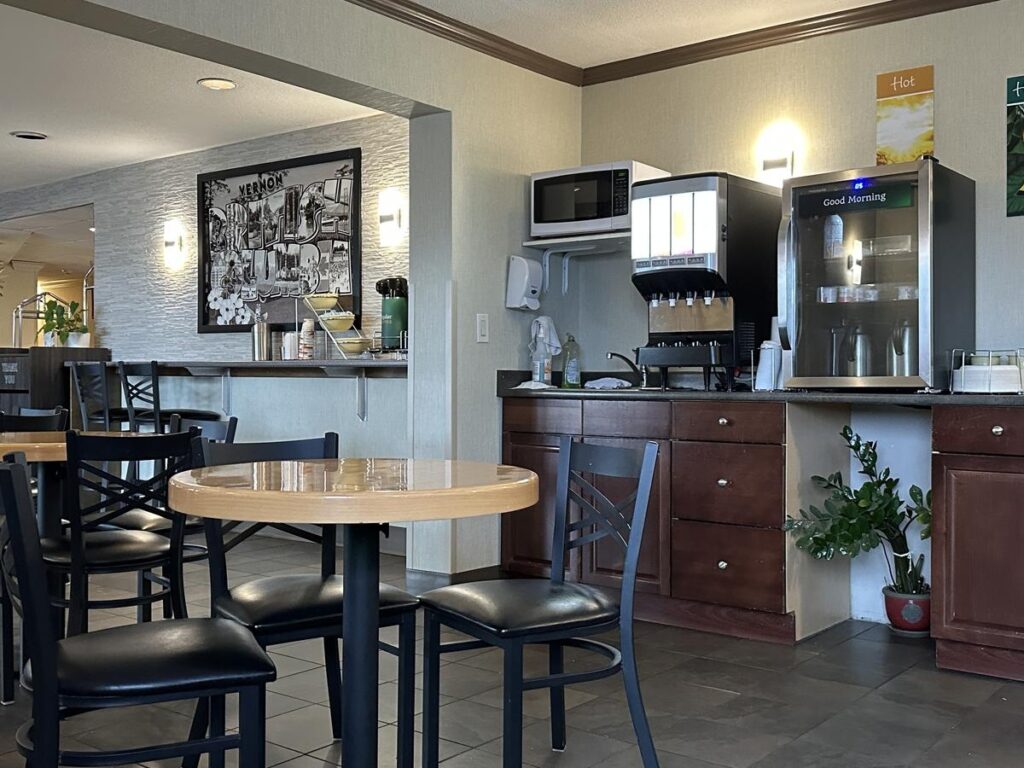 Dining area with wooden tables and black chairs, featuring a beverage station with a drink dispenser, coffee pots, microwave, and cup storage.