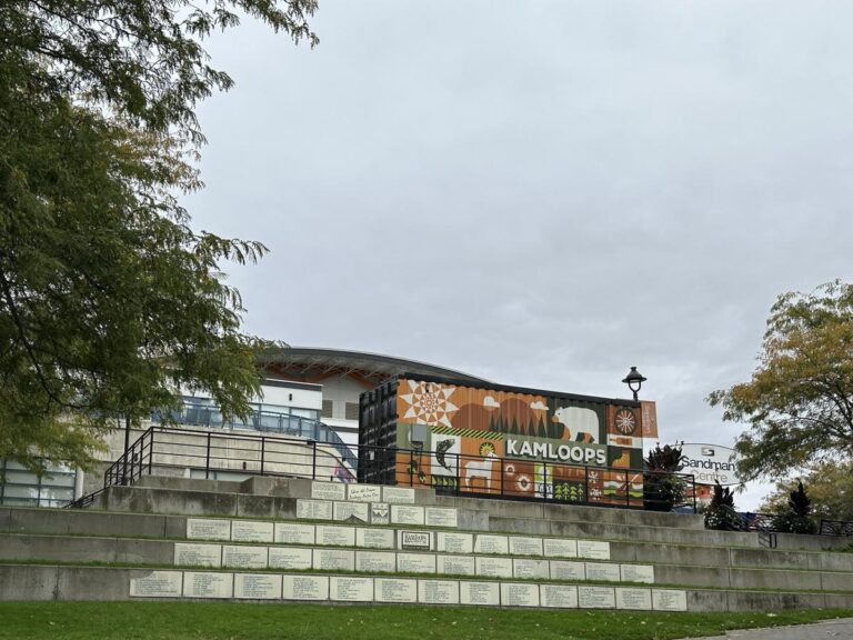 Colorful mural on a shipping container featuring stylized wildlife and landscape imagery with the word &ldquo;Kamloops&rdquo; in bold white letters. The mural sits in front of the Sandman Centre, framed by fall trees.