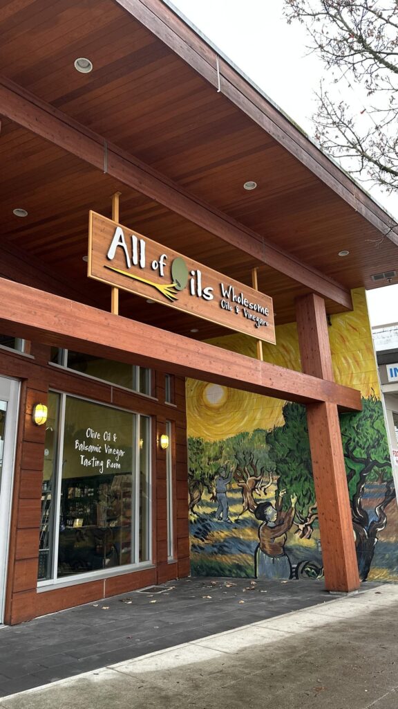 The storefront of All of Oils in Langley City, featuring a wooden sign and a large mural of people harvesting olives under a golden sun, with a glass window reading &ldquo;Olive Oil & Balsamic Vinegar Tasting Room.&rdquo;
