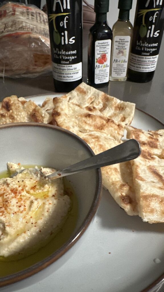A serving of creamy hummus drizzled with olive oil and sprinkled with chili flakes, surrounded by grilled flatbread on a plate, with All of Oils bottles and packaged Sahand bread in the background.