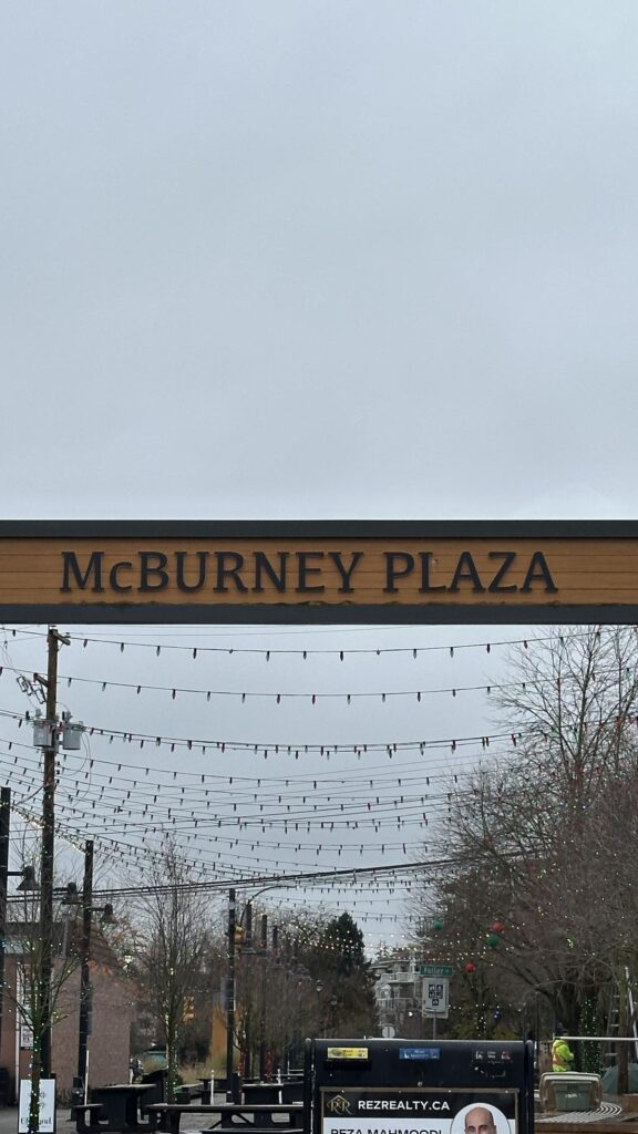 A wide shot of McBurney Plaza in Langley City under overcast skies, with strings of festive lights overhead and a wooden sign overhead reading &ldquo;McBurney Plaza.&rdquo;