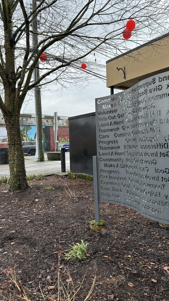 A steel sculpture installation features cut-out community words like &ldquo;Support,&rdquo; &ldquo;Volunteer,&rdquo; and &ldquo;Give Back,&rdquo; surrounded by bare trees and winter landscaping.