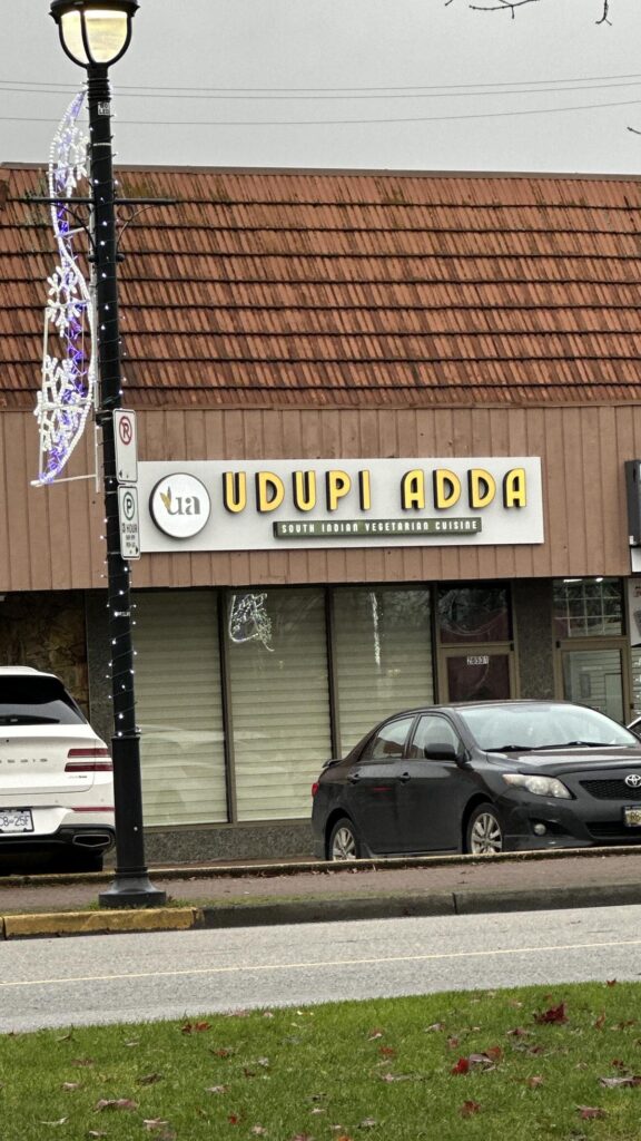 The storefront of Udupi Adda, a South Indian vegetarian restaurant in Langley, with a simple sign above two parked cars.