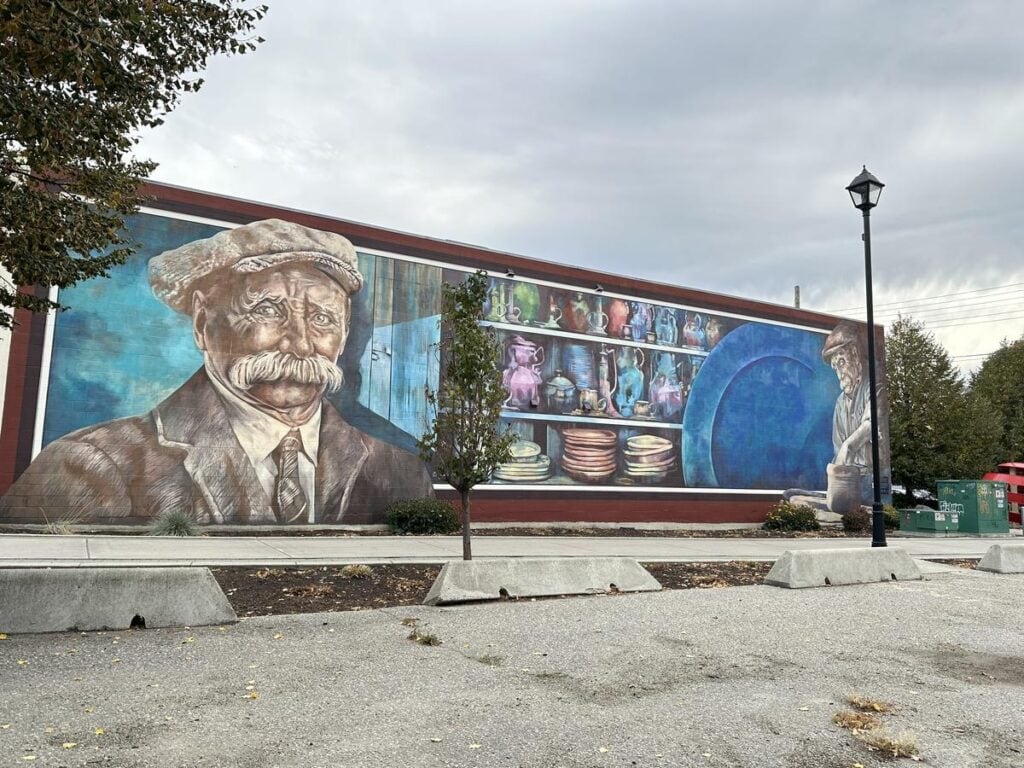 Full mural view of &ldquo;Pottery and Its Place&rdquo; showing two men, shelves filled with pottery, and a massive blue ceramic plate on the side of a downtown Vernon building.