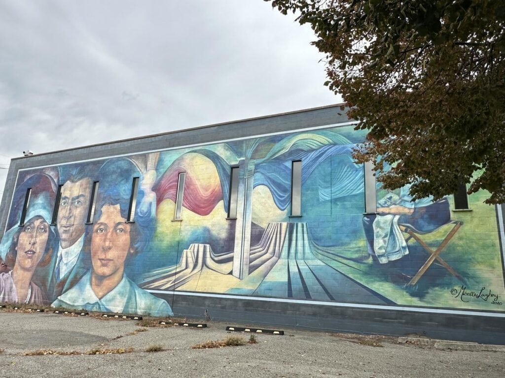 Mural titled &ldquo;The Captive&rdquo; Artist on a downtown Vernon wall, depicting stylised portraits of early 20th-century figures alongside surreal imagery of flowing fabric and a seated woman appearing to sew or mend.