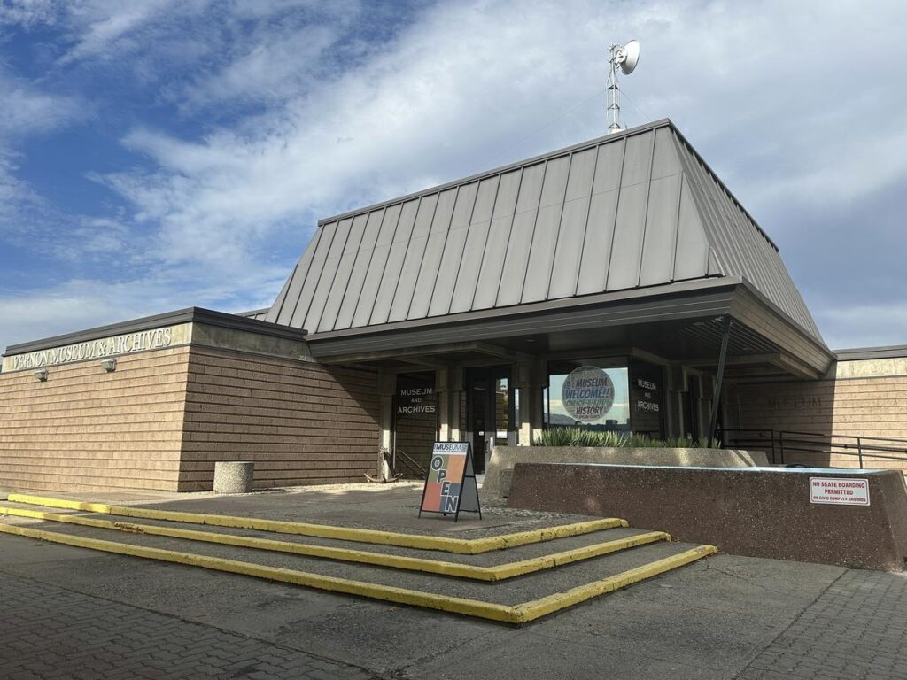 Wide view of the Vernon Museum & Archives entrance under a grey metal roof with an &ldquo;OPEN&rdquo; sign by the door and a blue sky above.