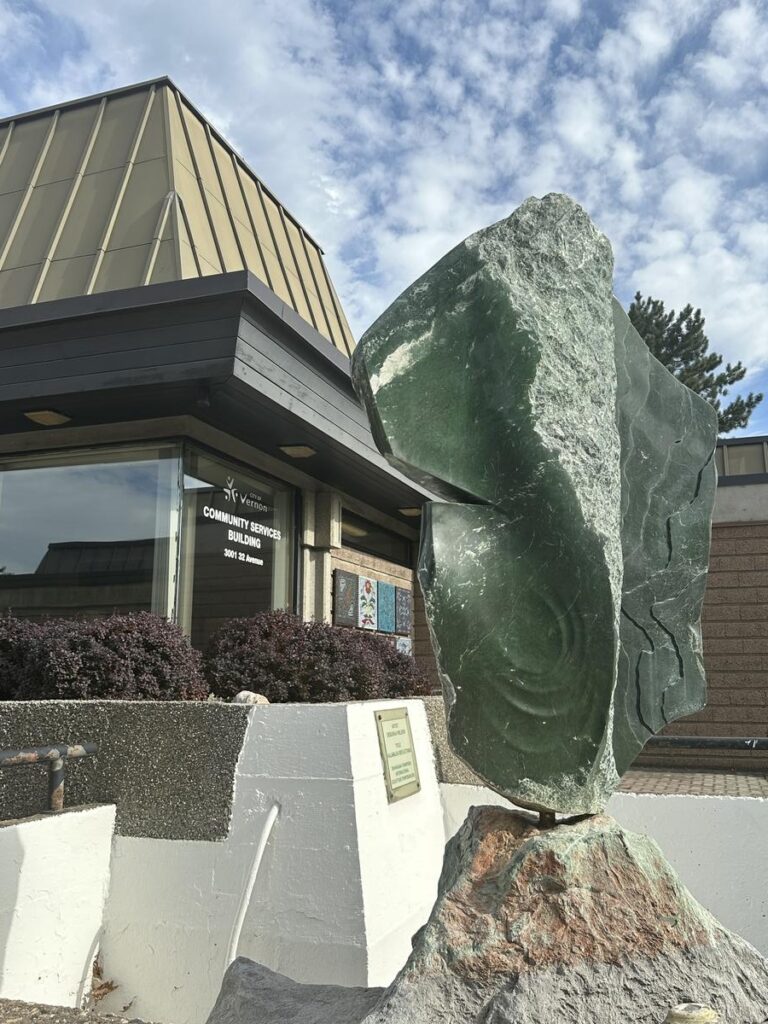 Abstract green jade sculpture on a white pedestal outside the Vernon Museum and Archives, with a partly cloudy sky behind.