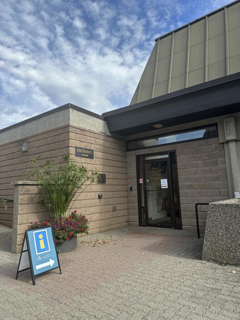 Main entrance to the Vernon Museum & Archives with a directional tourism info sign and potted flowers nearby.