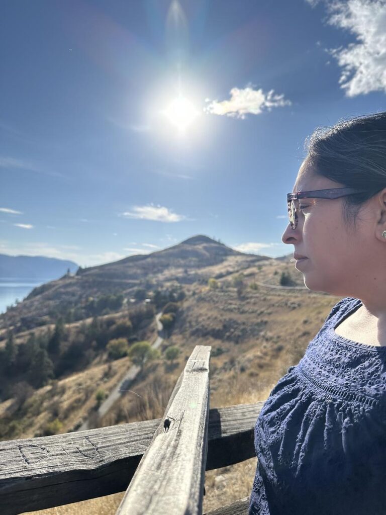 Tazim stands at a wooden railing gazing over sunlit hills and the brilliant blue of Kalamalka Lake, the midday sun shining brightly.