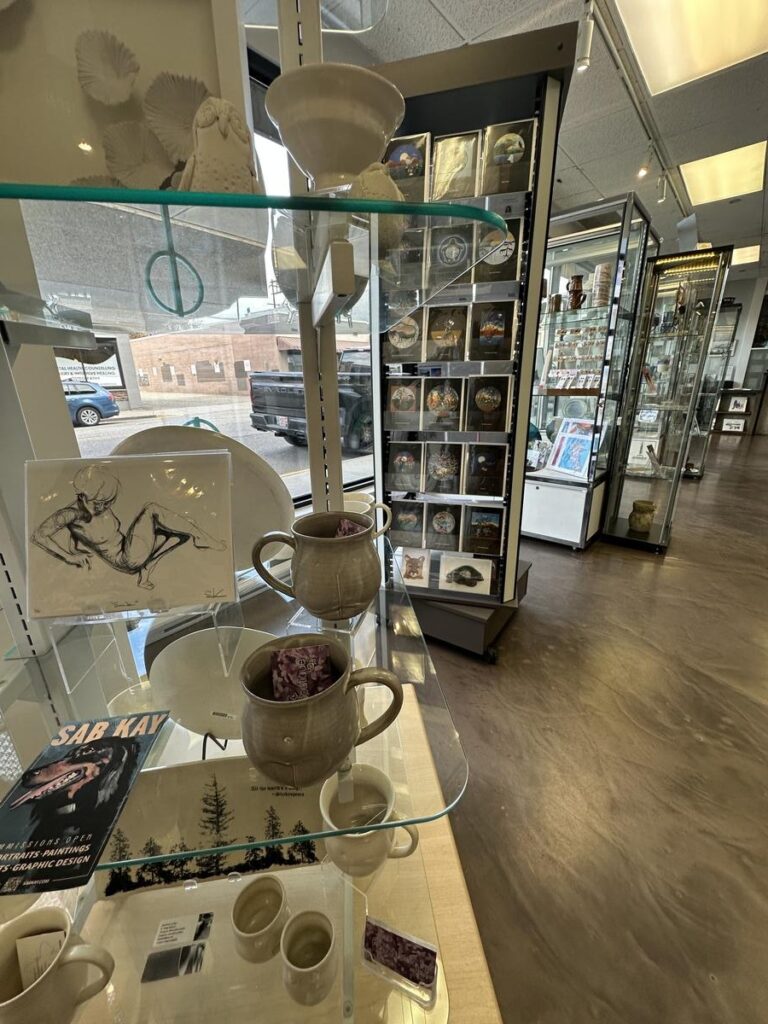 Shelves of handmade pottery, cards, and small artworks in the Vernon Public Art Gallery&rsquo;s gift shop, with display cases and a view of the street outside.