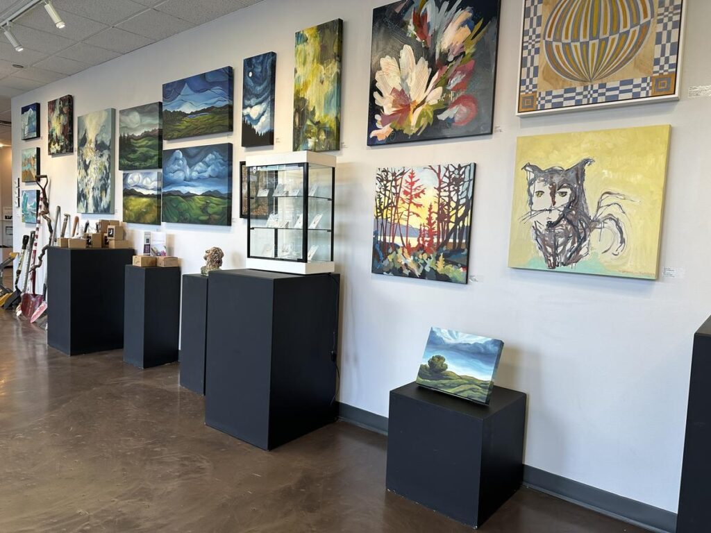 A well-lit gallery wall at the Vernon Public Art Gallery displaying a mix of landscape, abstract, and figurative paintings, with display stands below.