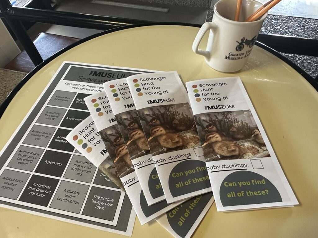 A set of scavenger hunt brochures and a bingo-style activity sheet sit on a round table beside a mug with pencils, promoting a kids' activity at the Vernon Museum.