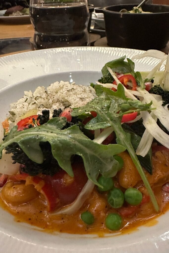 A plant-based dish at Atlas Steak + Fish, Delta Hotels Burnaby Conference Centre, featuring a tomato-based vegetable stew with peas, cherry tomatoes, kale, fennel, and arugula, served with herbed rice on a white plate, Burnaby, BC.