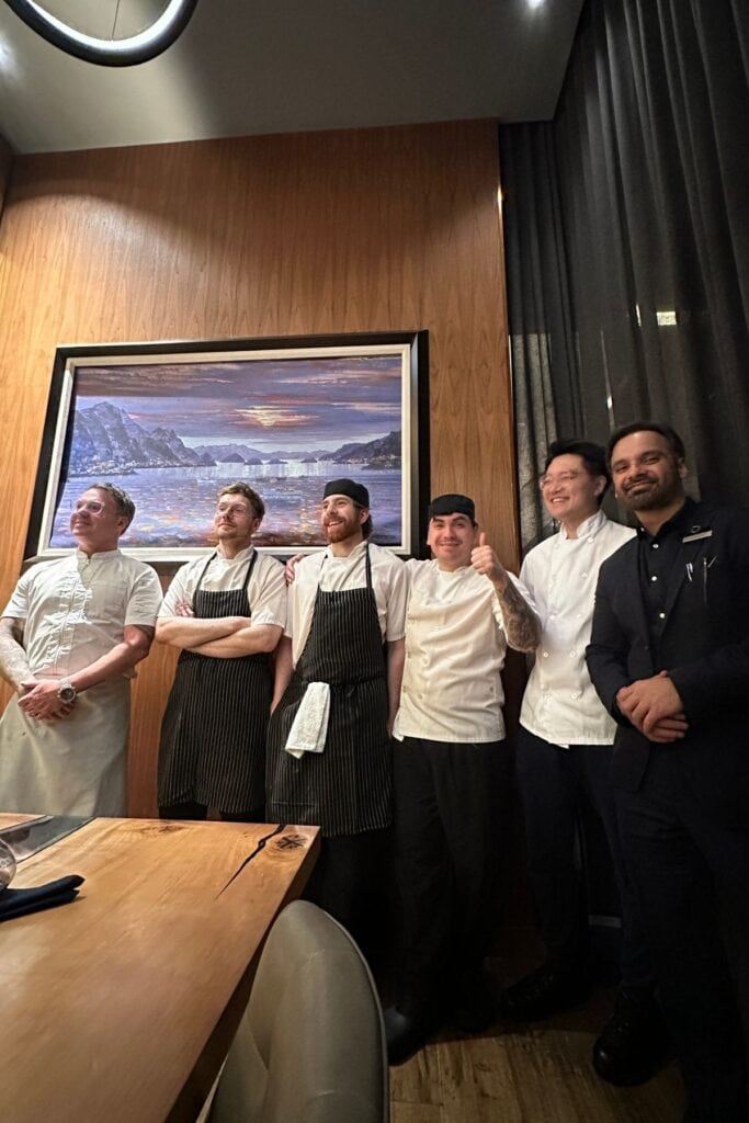 Chef Spencer Watts and the kitchen and front-of-house team at Atlas Steak + Fish inside the Delta Hotels Burnaby Conference Centre, standing in front of a landscape painting in the restaurant dining room, Burnaby, BC.