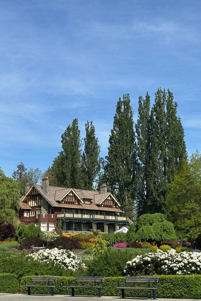 The Burnaby Art Gallery, a Tudor-style heritage building surrounded by blooming white and pink gardens, with tall poplar trees and a blue sky, viewed from the park benches at Deer Lake Park, Burnaby, BC.