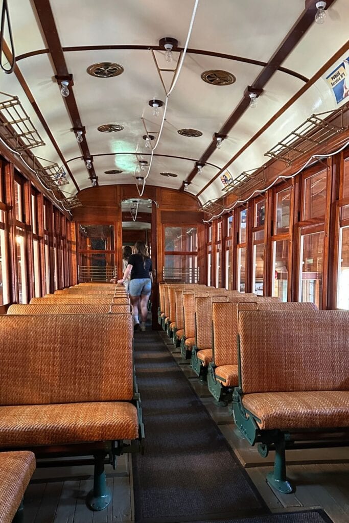 The restored interior of Interurban Tram 1223 at Burnaby Village Museum, showing rows of woven rattan seats, polished wood panelling, a curved white ceiling with period globe lights, and a visitor walking toward the far end, Burnaby, BC.