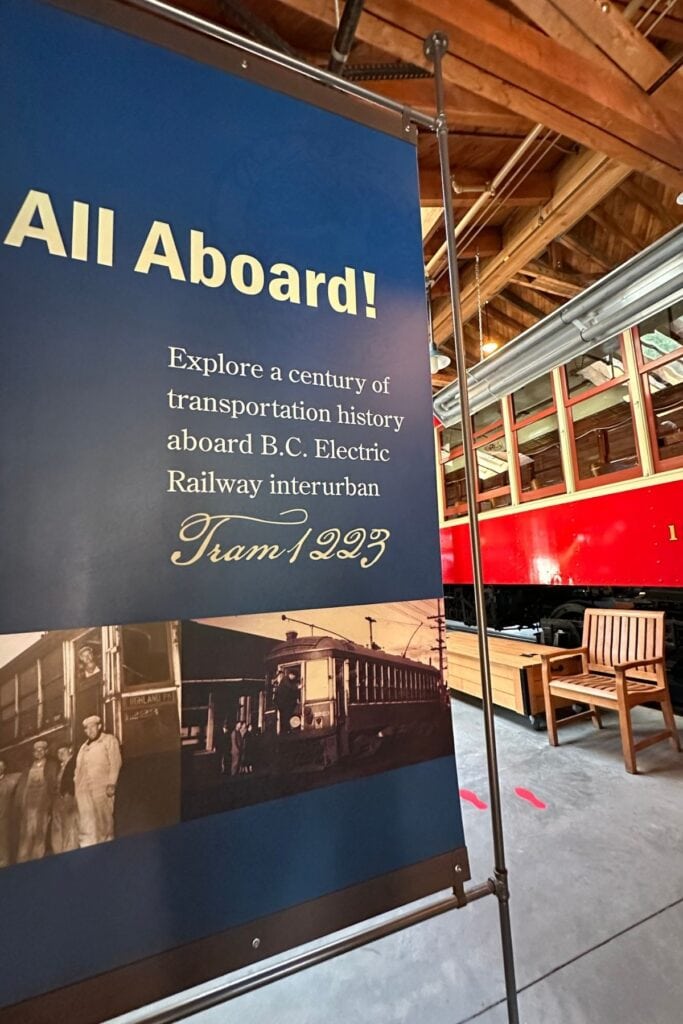 An All Aboard exhibition sign in front of Interurban Tram 1223 at Burnaby Village Museum, inviting visitors to explore a century of transportation history aboard the B.C. Electric Railway interurban tram, Burnaby, BC.