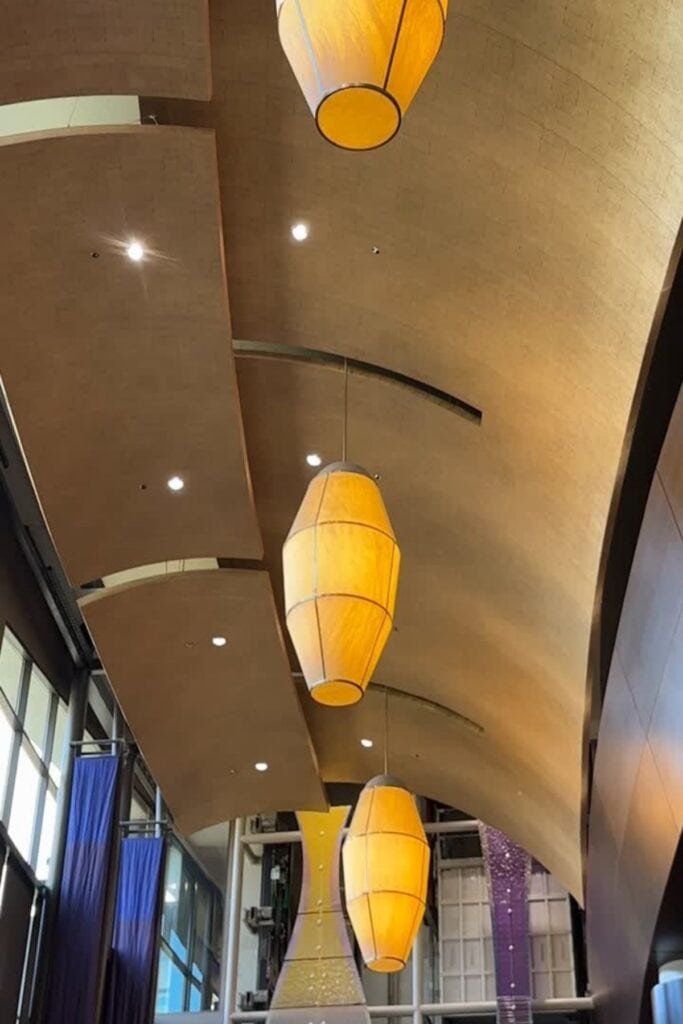 The lobby ceiling of the Delta Hotels Burnaby Conference Centre, featuring large amber pendant lantern lights hanging from a curved wooden ceiling, with blue and purple decorative elements from the connected Grand Villa Casino visible below, Burnaby, BC.