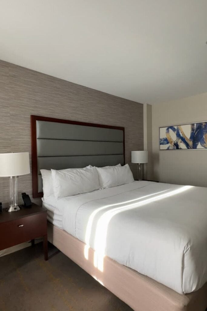 A clean, modern king room at the Delta Hotels Burnaby Conference Centre, with a grey upholstered headboard, white bedding, dark wood bedside tables, bedside lamps, textured wallpaper, and an abstract artwork on the wall, Burnaby, BC.