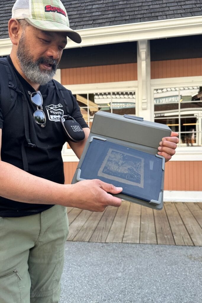 Erwin Rosales from Storyboard Experiences holding a tablet showing a production still at Burnaby Village Museum, standing in front of the 1920s main street storefronts, Burnaby, BC.